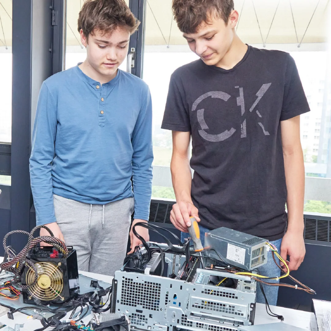 Zwei Jugendliche stehen an einem geöffneten Desktop-PC auf einem Tisch; einer benutzt einen Schraubenzieher, neben ihnen liegen Netzteile, Kabel und Hardwarekomponenten, sie reparieren oder bauen den Computer.