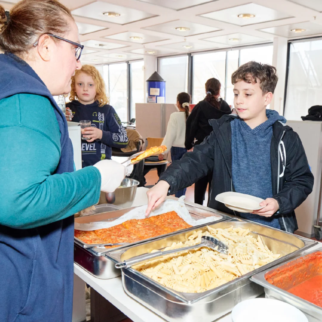 Frau serviert einem Jungen an einer Essensausgabe ein Stück Auflauf; Tabletts mit Pasta und Tomatensauce stehen bereit, weitere Kinder warten in der Schlange.